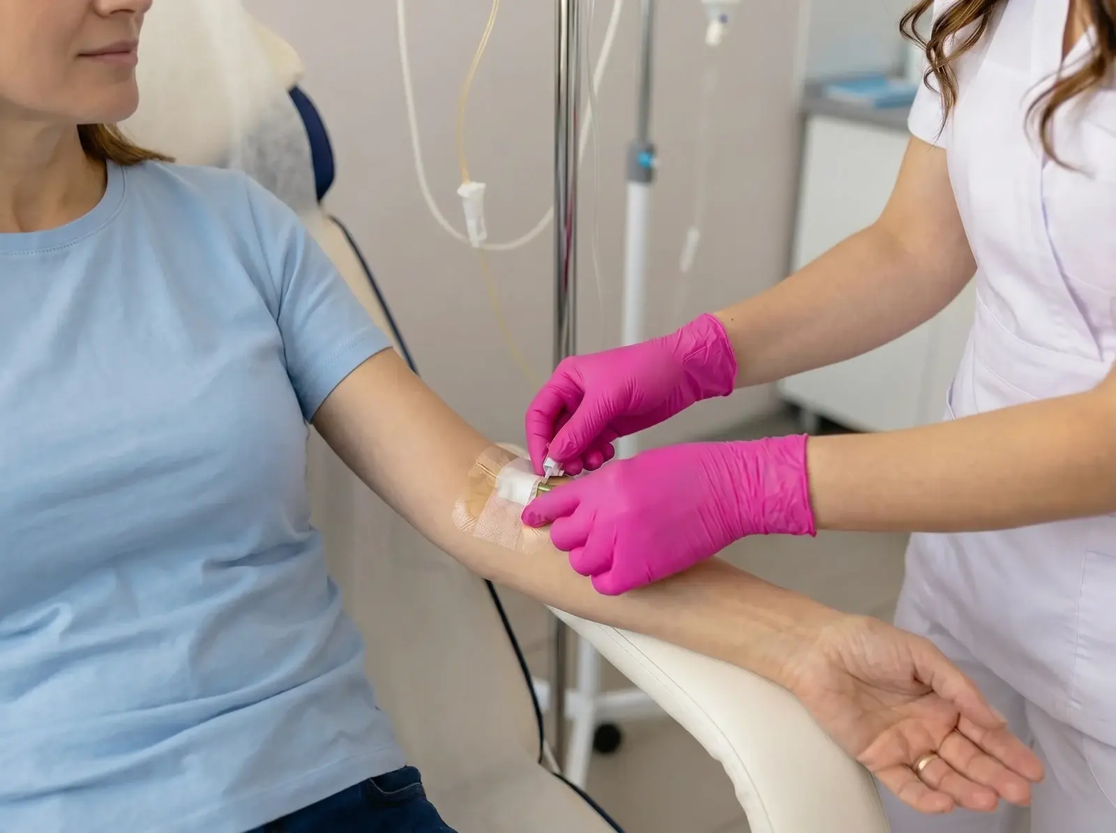 Nurse in a white uniform and bright pink gloves pressing down the tape and cap of an IV cannula in the forearm of a seated woman in a light blue t-shirt, with IV tubing and a drip stand behind them